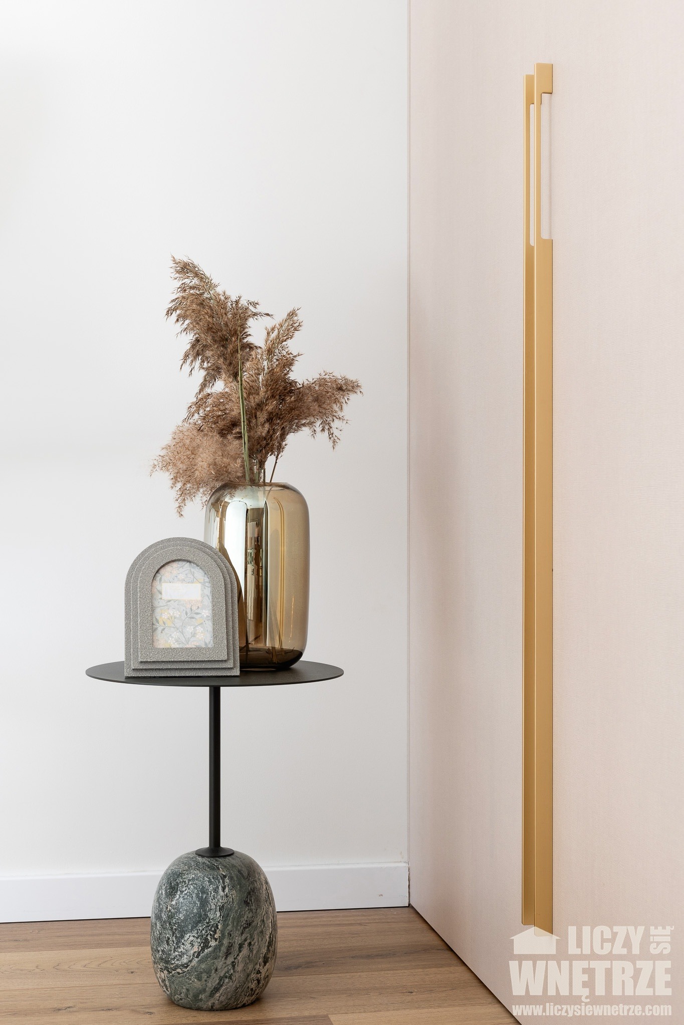 Dekoracyjny stolik z marmurową podstawą, złotym wazonem z trawami i ramką na zdjęcia, ustawiony przy ścianie z długim, złotym uchwytem.