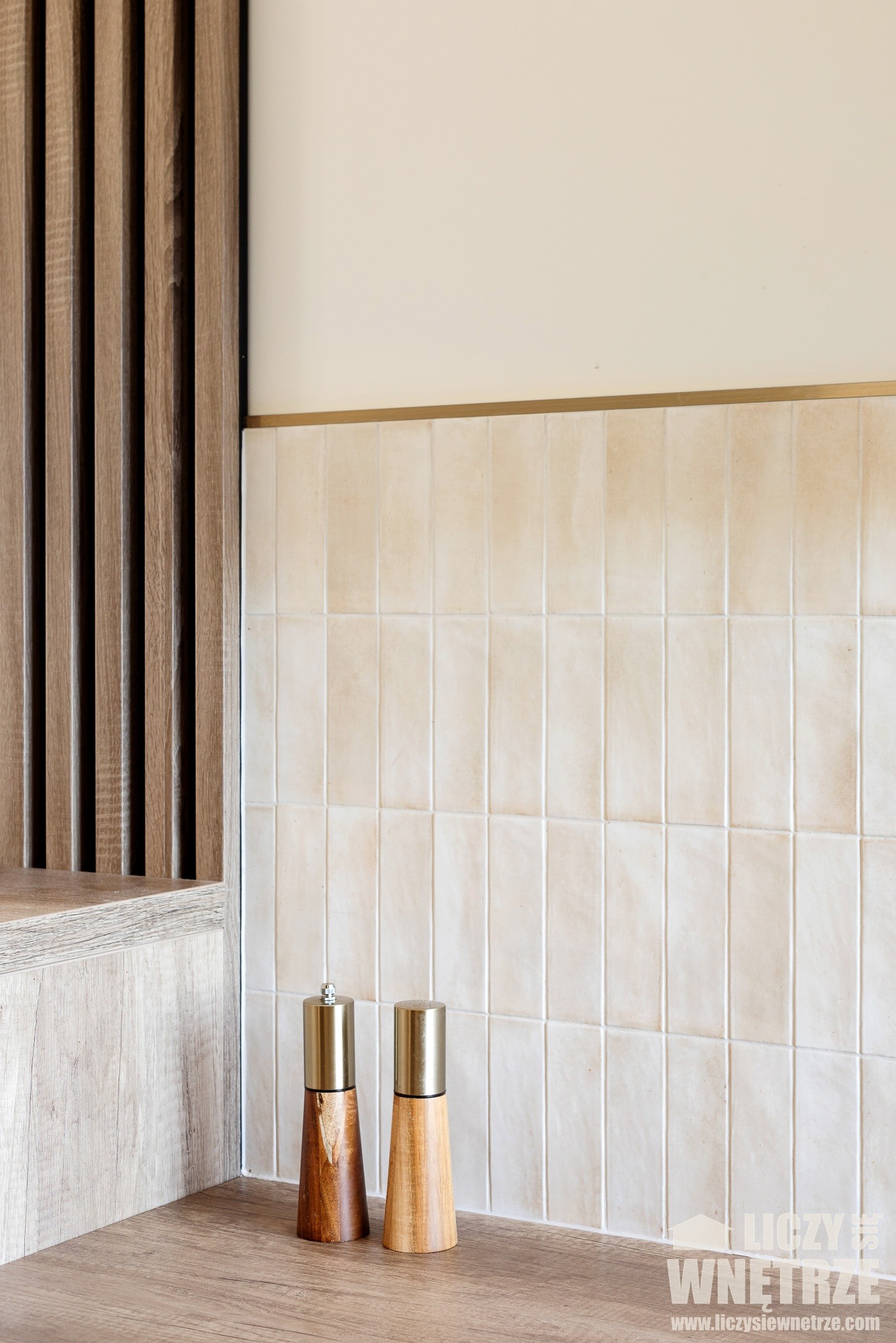 Kuchnia w odcieniach beżu z drewnianymi akcentami: pionowe lamele, kremowe płytki i przyprawniki. Minimalistyczny design, ciepłe oświetlenie.