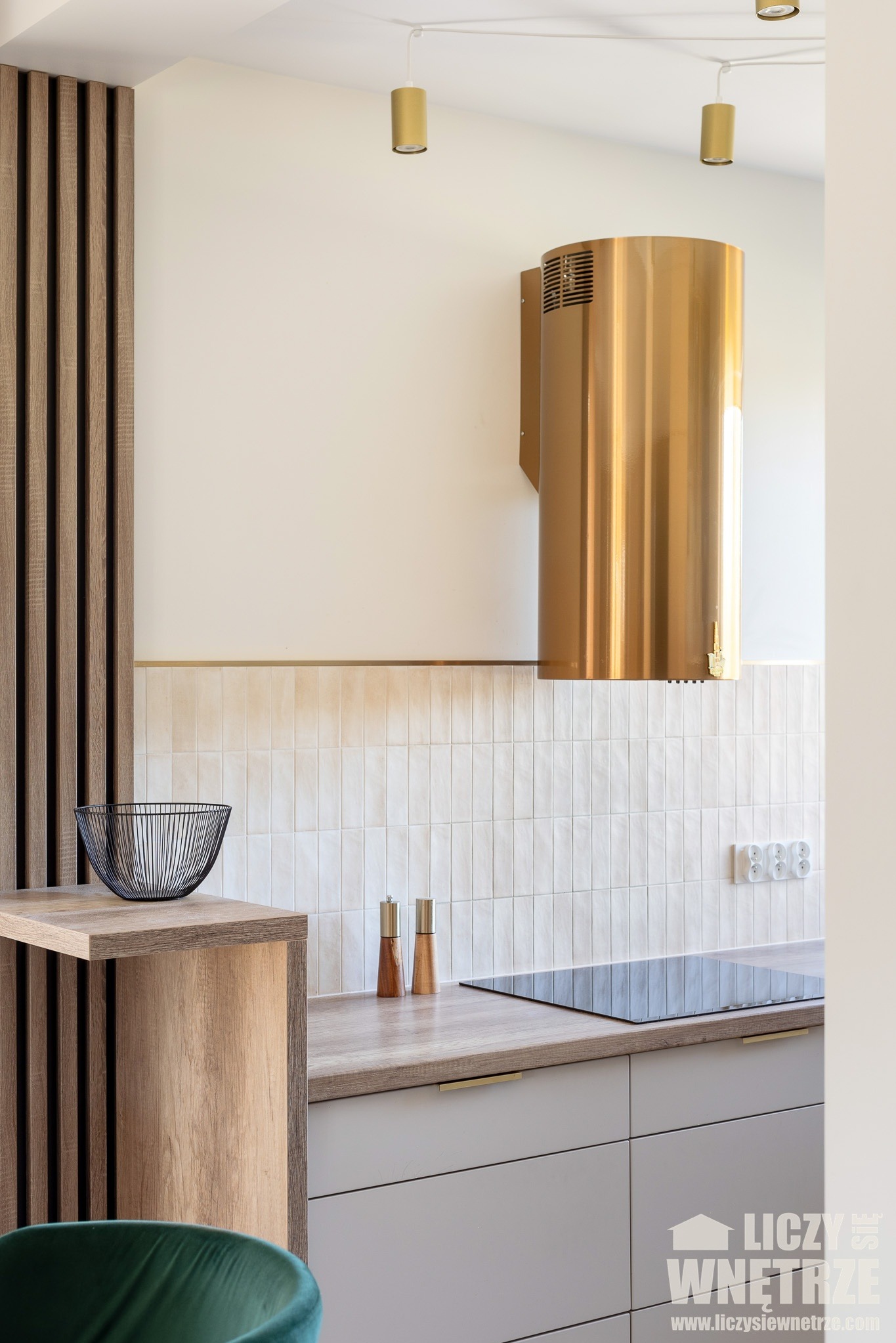 Nowoczesna kuchnia z złotym okapem cylindrycznym, drewnianą ścianką z listew i blatem w kolorze drewna. Minimalistyczny design, płytki w odcieniach beżu.