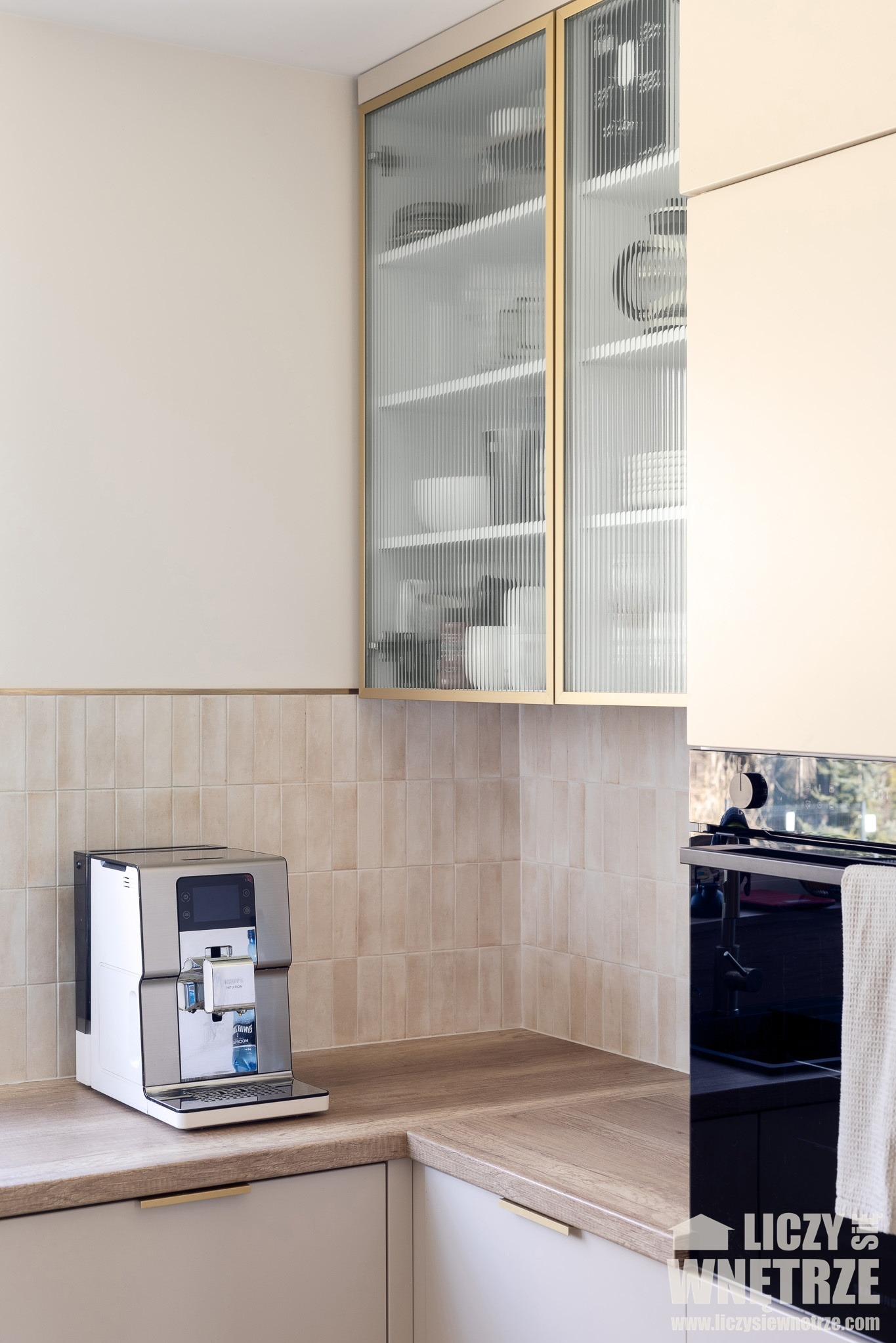 Kuchenna zabudowa z ryflowanymi szklanymi frontami szafek, beżowe płytki na ścianie i ekspres do kawy na blacie. Minimalistyczny design, ciepłe oświetlenie.