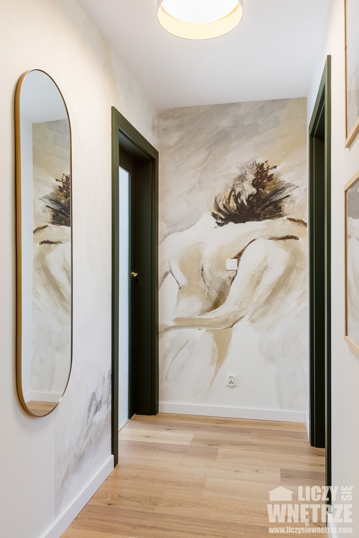 Korytarz z lustrem w złotej ramie i malowidłem na ścianie w odcieniach beżu i brązu. Podłoga drewniana, drzwi w ciemnej zieleni.