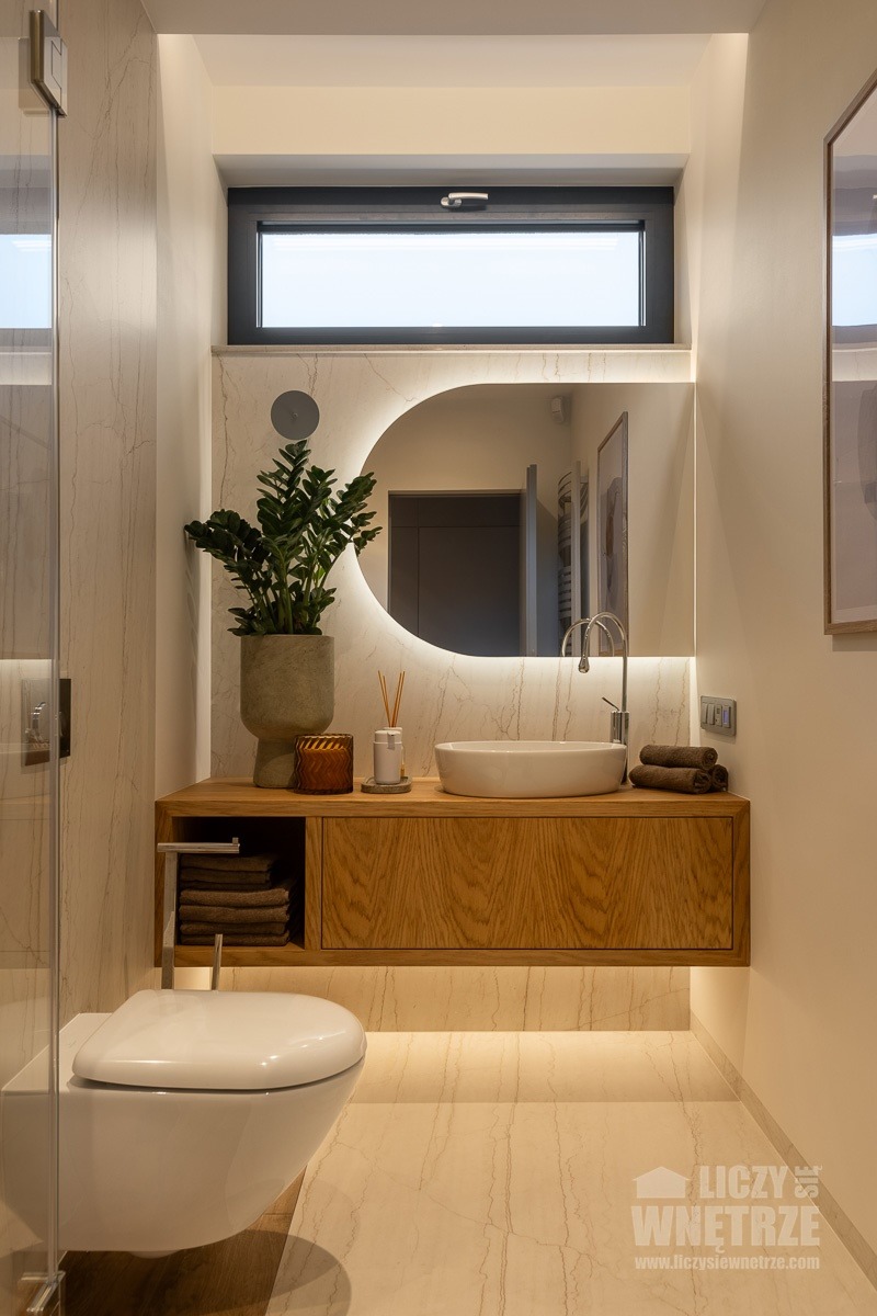 Nowoczesna łazienka z podświetlanym lustrem, drewnianą szafką pod umywalką i wiszącą toaletą. Na blacie roślina i akcesoria. Neutralna kolorystyka z akcentami drewna.