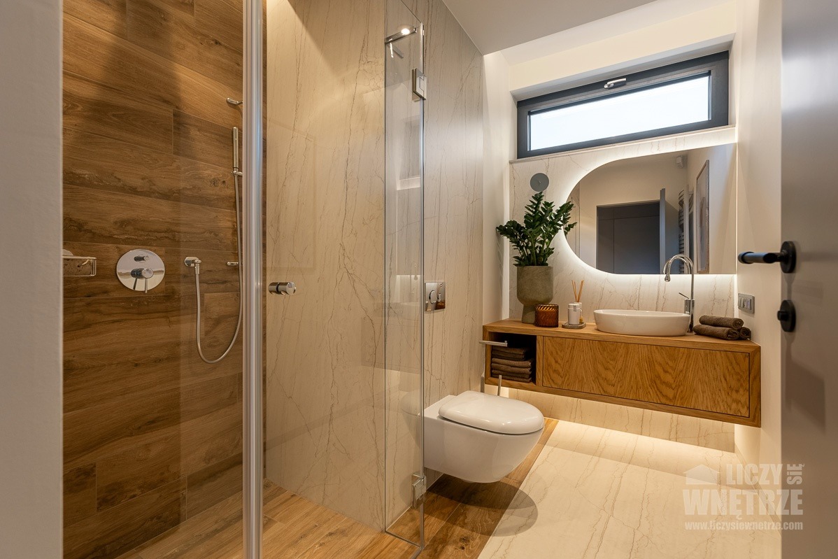 Nowoczesna łazienka z drewnianymi akcentami: kabina prysznicowa, podświetlane lustro, wisząca toaleta i szafka pod umywalką. Minimalistyczny design.