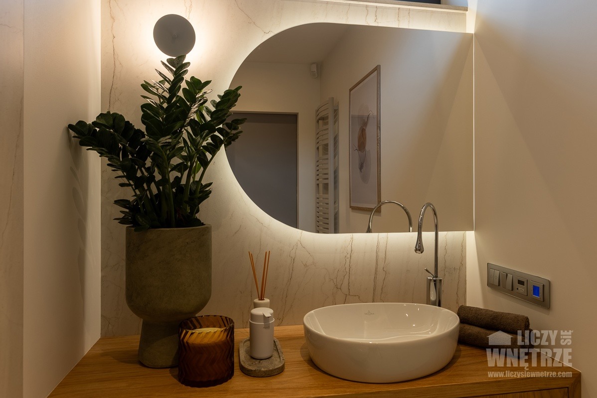 Elegancka łazienka z okrągłym lustrem podświetlanym LED, umywalką nablatową i zielonym kwiatem. Marmurowe wykończenie ściany i drewniany blat dodają ciepła.