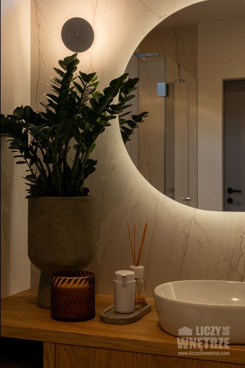 Stylowa aranżacja łazienki: okrągłe lustro z podświetleniem LED, umywalka nablatowa, roślina w doniczce i dekoracje na blacie z drewna. Minimalistyczny design.