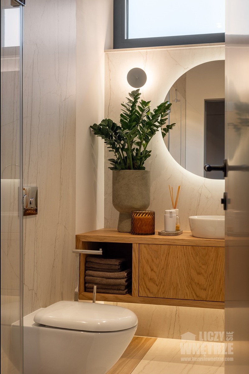 Nowoczesna łazienka z drewnianą szafką pod umywalką, okrągłym lustrem z podświetleniem LED i rośliną w doniczce. Minimalistyczny design z akcentami natury.