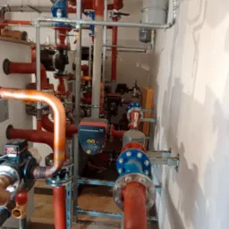 Złożona instalacja grzewcza z rur miedzianych i stalowych, z pompami obiegowymi Grundfos i zaworami, zamontowana w pomieszczeniu technicznym przy białej ścianie.
