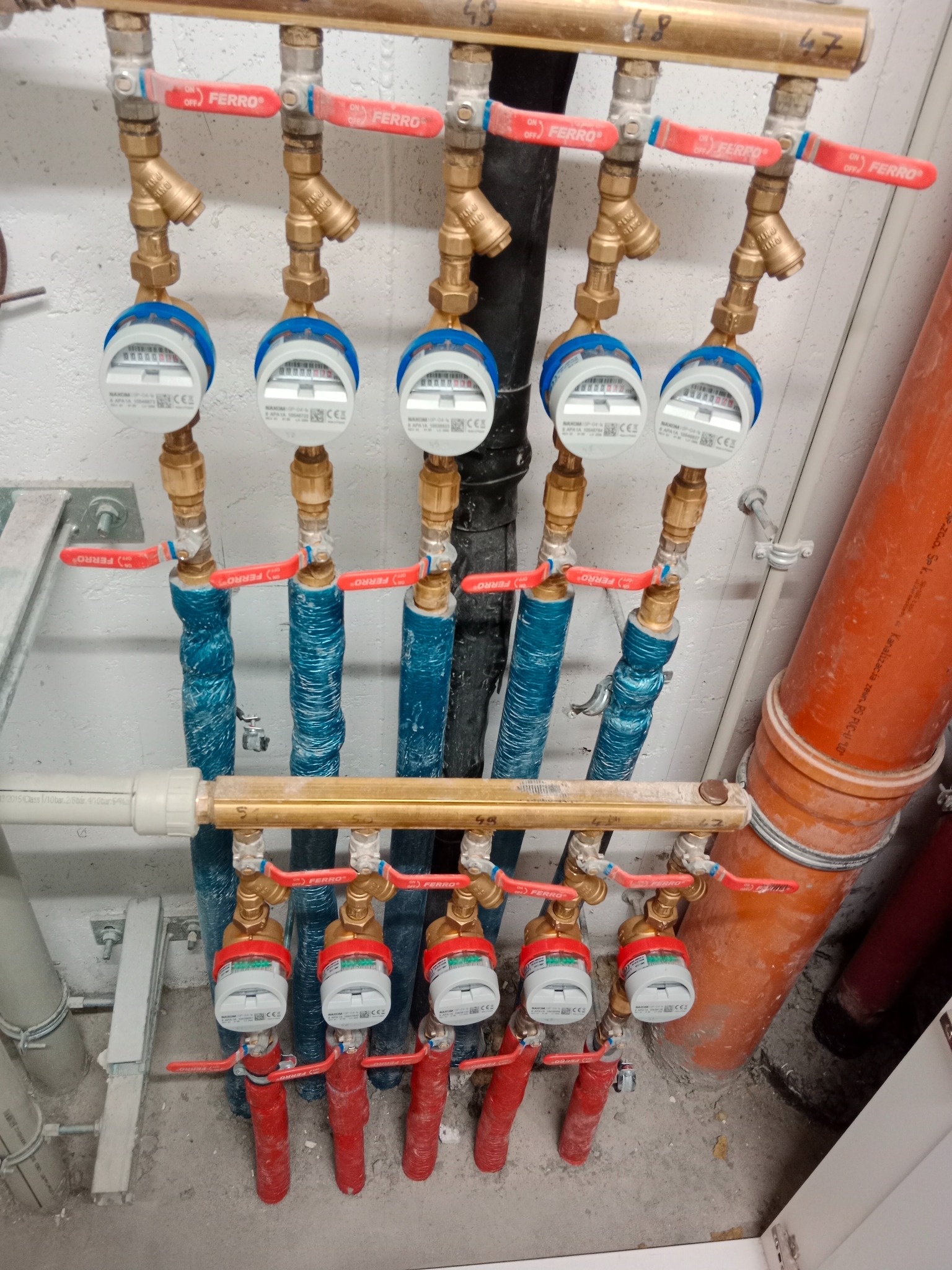 Rozdzielacz instalacji wodnej z zaworami FERRO i licznikami, rury zaizolowane na niebiesko i czerwono, widoczne numery 45, 46, 47, 48.