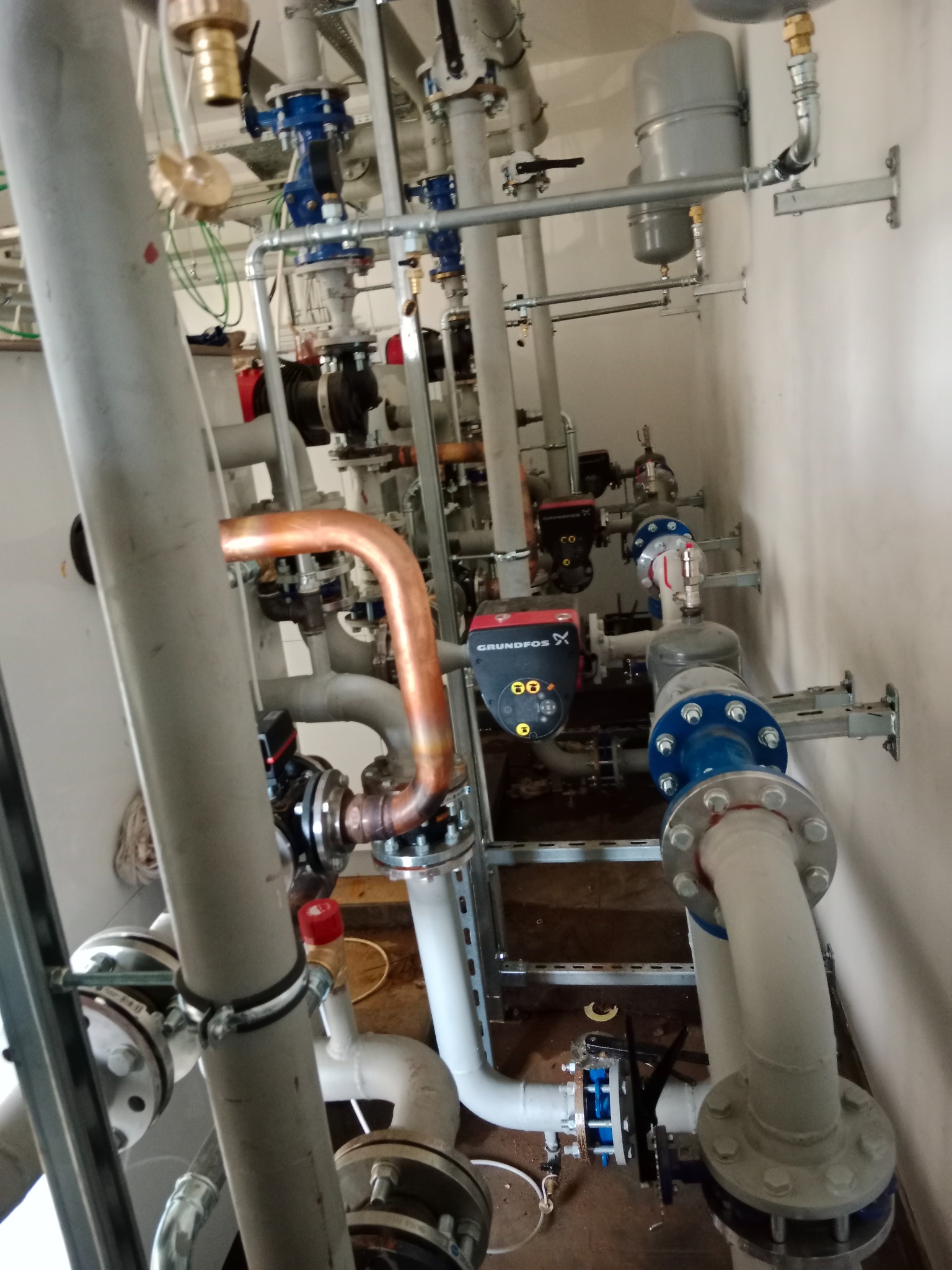 Złożona instalacja grzewcza w kotłowni z rurami, zaworami, pompą Grundfos i zbiornikami wyrównawczymi. Widok z dołu, oświetlenie naturalne.