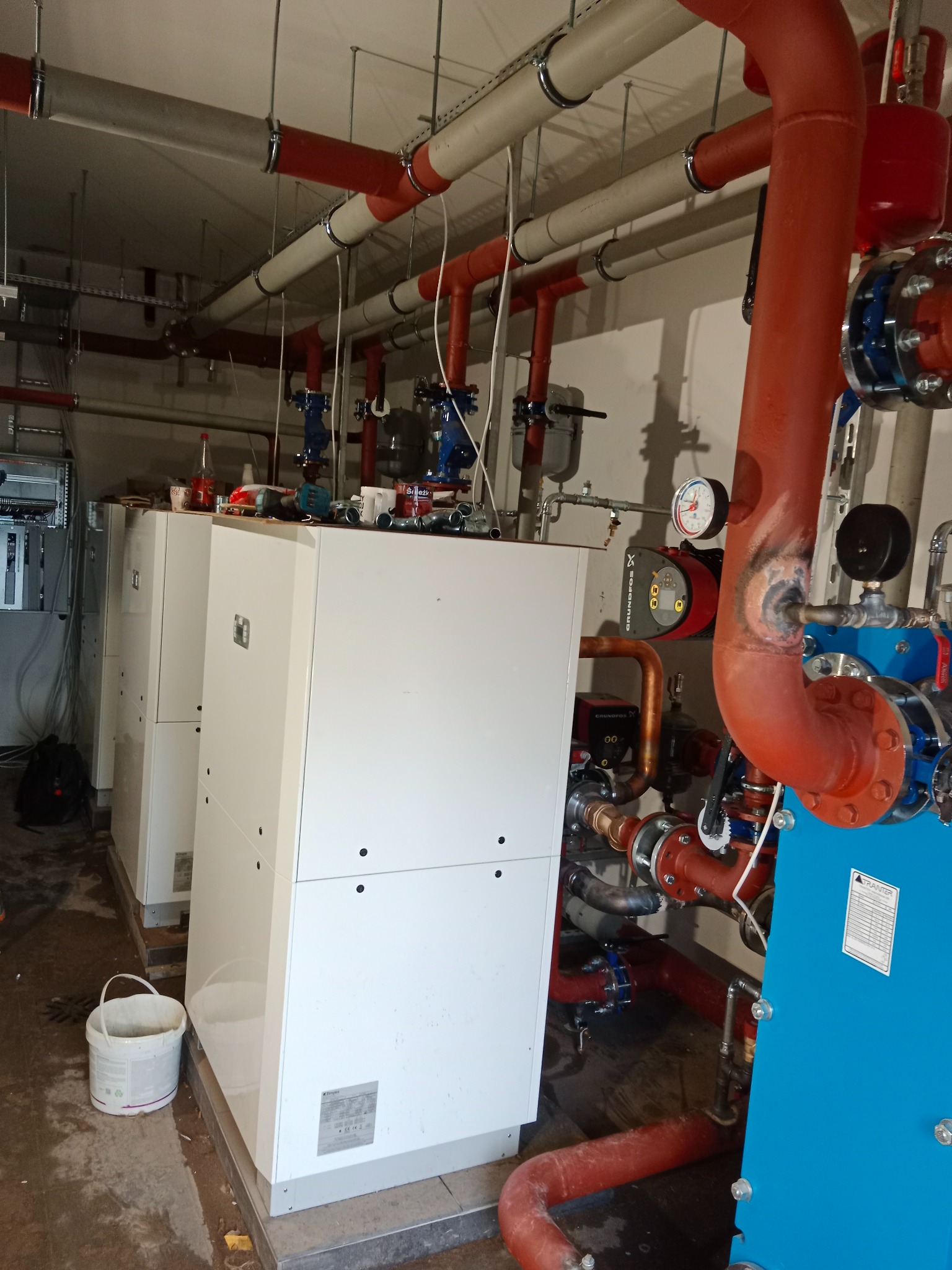 Kotłownia z trzema białymi kotłami i systemem rur w kolorze czerwonym, niebieskim i miedzianym. Widoczne manometry i pompy obiegowe. Na podłodze stoi wiadro.