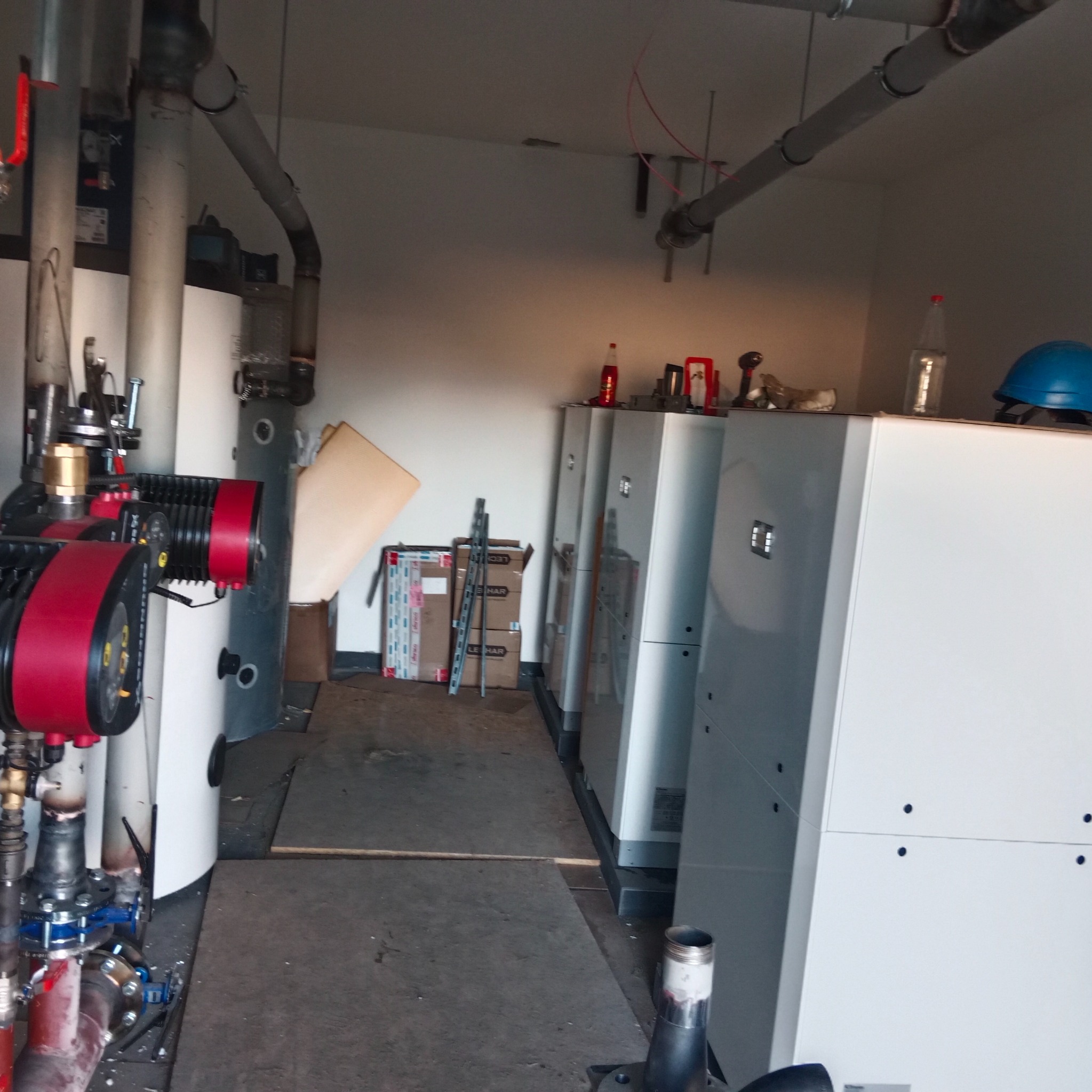 Instalacja grzewcza w kotłowni: białe piece CO, czerwona pompa obiegowa, rury. Na piecach leżą narzędzia i kask. Widok na podłogę i ścianę z rurami.