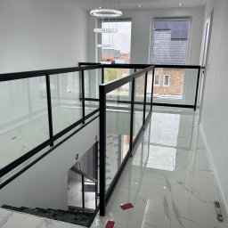 Hoff-Metall - Nowoczesna, czarna balustrada szklana na schodach, z widokiem na okno i elegancki żyrandol. Białe ściany i marmurowe płytki podłogowe tworzą minimalistyczny design.