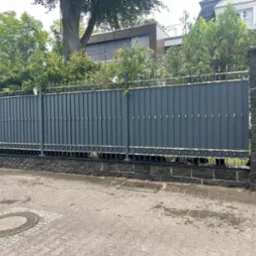 Nowoczesna, grafitowa brama wjazdowa z dekoracyjnymi elementami w kolorze złota, osadzona na kamiennych słupkach i podmurówce, widok z zewnątrz posesji.