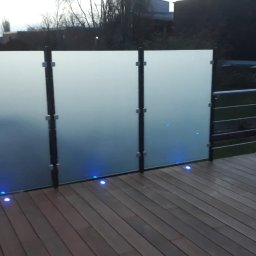 Hoff-Metall - Szklana balustrada balkonowa z podświetleniem LED wbudowanym w drewnianą podłogę. Matowe szkło zapewnia prywatność, a metalowe słupki stabilność.