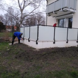 Hoff-Metall - Montaż balustrady szklanej na tarasie: pracownik w niebieskiej bluzie mocuje element, w tle balkon i osoba w czerwonej bluzie. Widok z zewnątrz, w tle drzewa.