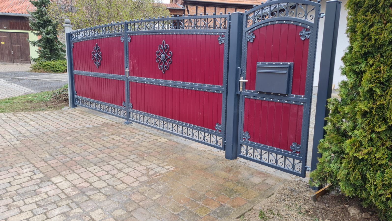 Czerwono-szara brama wjazdowa z dekoracyjnymi elementami, furtka z skrzynką na listy. Kostka brukowa na podjeździe, krzew iglasty po prawej.