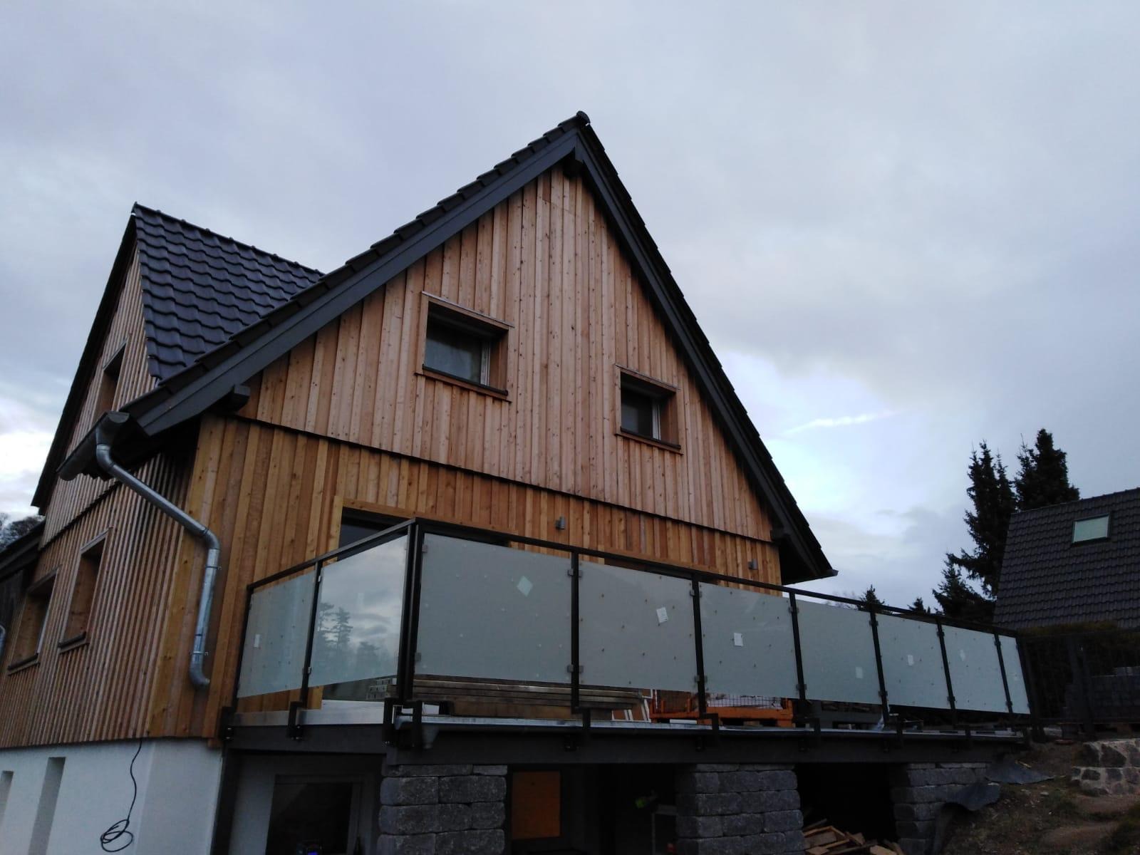 Drewniany dom z balkonem i szklanymi balustradami. Nowoczesny design, widok z dołu, pochmurne niebo w tle. Charakterystyczna architektura budynku.