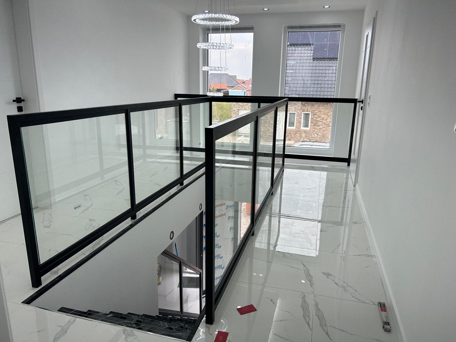 Nowoczesna, czarna balustrada szklana na schodach, z widokiem na okno i elegancki żyrandol. Białe ściany i marmurowe płytki podłogowe tworzą minimalistyczny design.