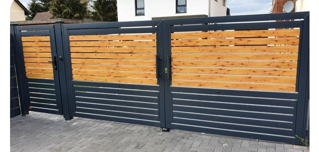 Nowoczesna brama wjazdowa z szarymi, metalowymi panelami i drewnianymi wstawkami. Widoczne poziome deski i metalowa konstrukcja. Brama na posesję.