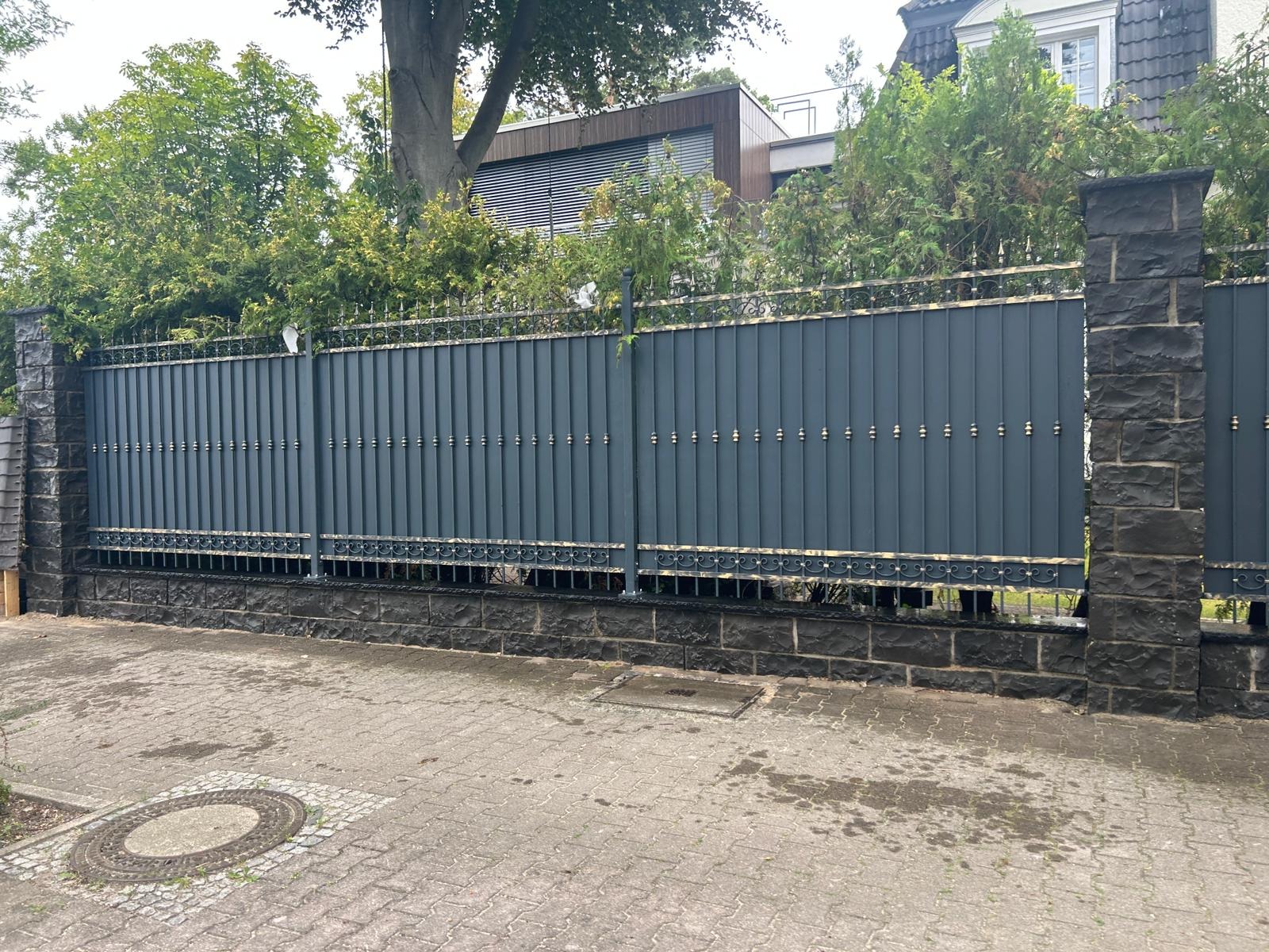 Nowoczesna, grafitowa brama wjazdowa z dekoracyjnymi elementami w kolorze złota, osadzona na kamiennych słupkach i podmurówce, widok z zewnątrz posesji.