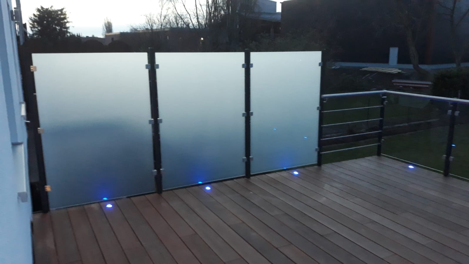 Szklana balustrada balkonowa z podświetleniem LED wbudowanym w drewnianą podłogę. Matowe szkło zapewnia prywatność, a metalowe słupki stabilność.