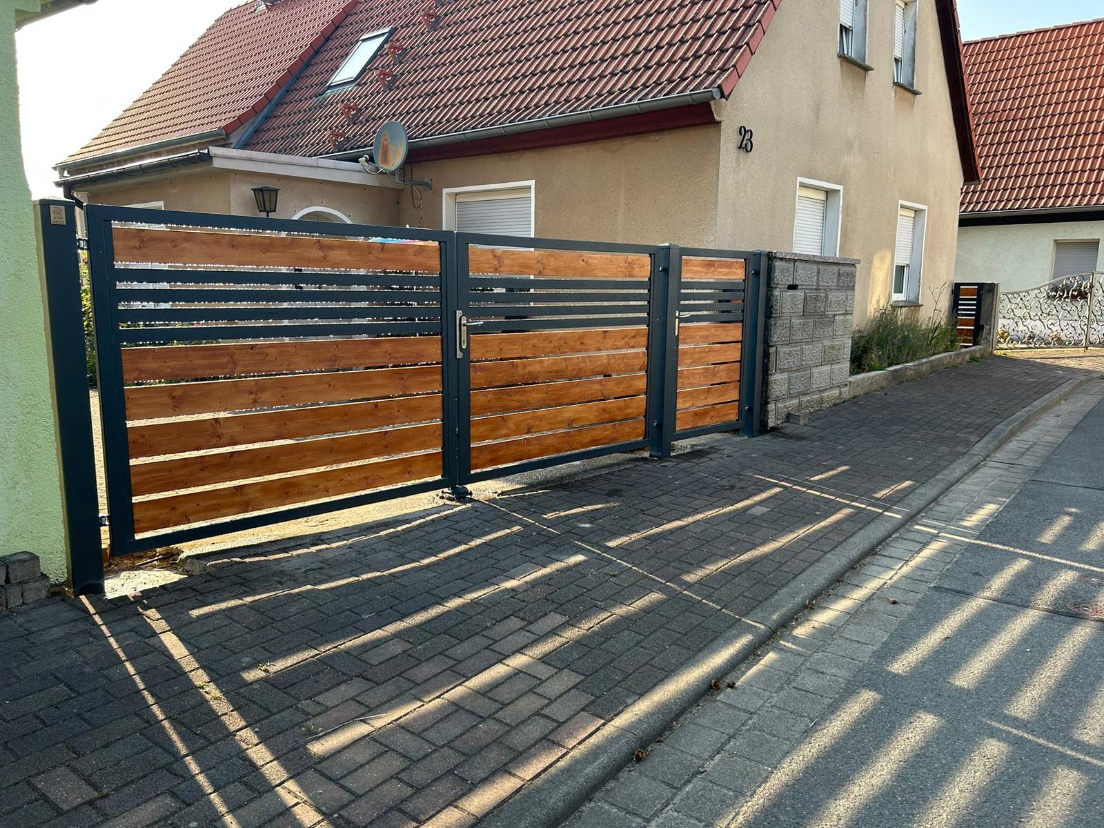 Nowoczesna brama posesyjna z drewnianymi panelami i metalową ramą, oświetlona promieniami słońca rzucającymi cienie na brukowaną nawierzchnię.