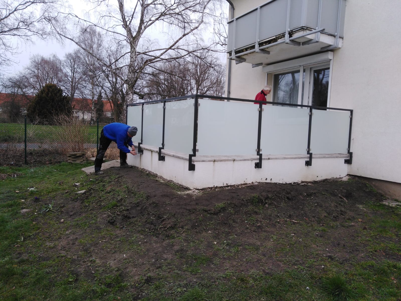 Montaż balustrady szklanej na tarasie: pracownik w niebieskiej bluzie mocuje element, w tle balkon i osoba w czerwonej bluzie. Widok z zewnątrz, w tle drzewa.