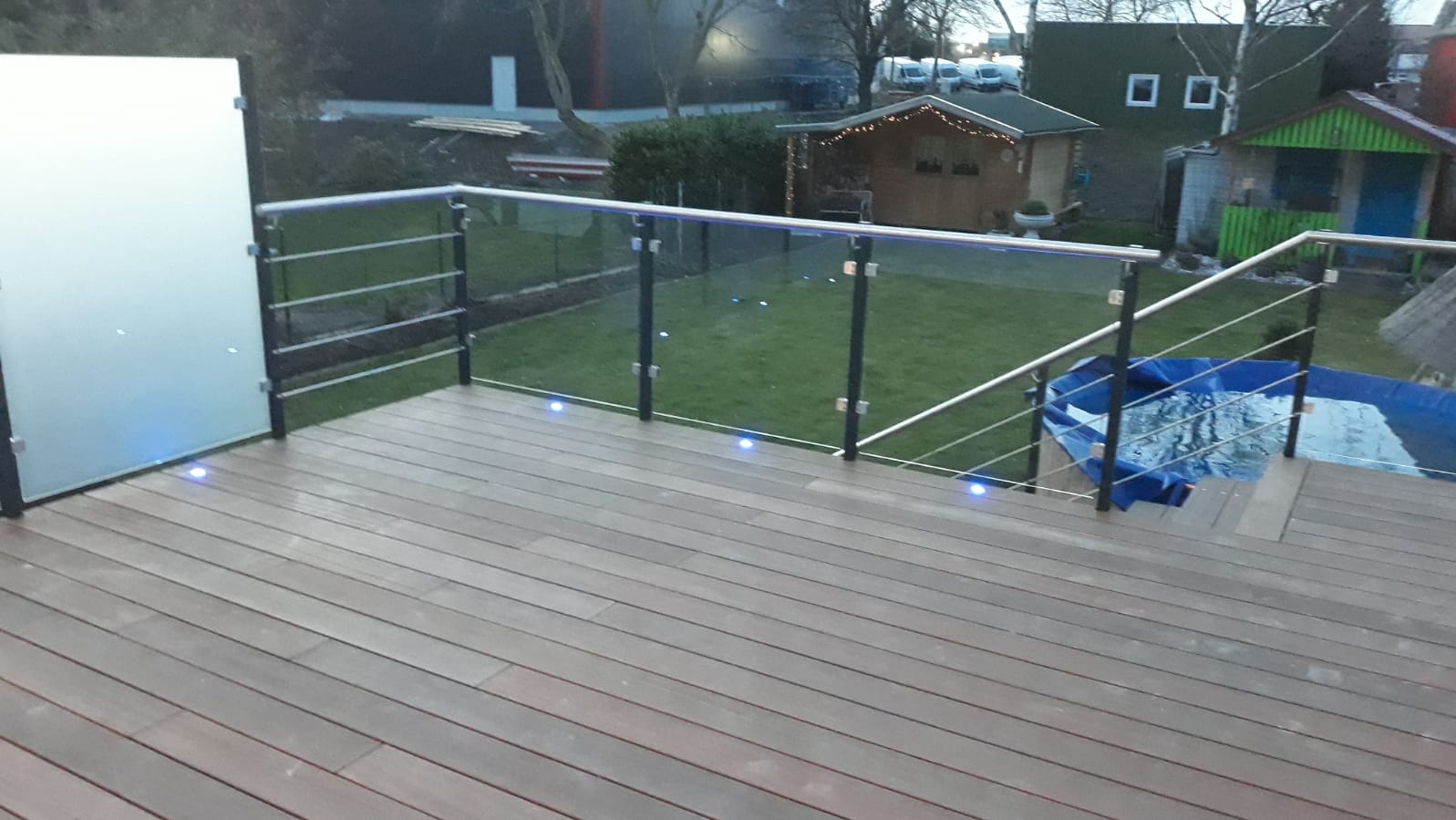 Szklana balustrada z oświetleniem LED na tarasie z desek kompozytowych, widok na ogród z domkami narzędziowymi i basenem. Nowoczesny design.