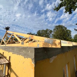 NOX INTERNATIONAL SPÓŁKA Z OGRANICZONĄ ODPOWIEDZIALNOŚCIĄ - Wznoszenie drewnianej więźby dachowej na budynku z żółtą elewacją, widoczne elementy konstrukcyjne i metalowe łączniki, błękitne niebo w tle.