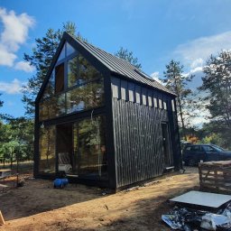 NOX INTERNATIONAL SPÓŁKA Z OGRANICZONĄ ODPOWIEDZIALNOŚCIĄ - Nowoczesny, czarny dom modułowy z dużymi przeszkleniami, otoczony lasem. Widoczne elementy budowy i samochody w tle. Ujęcie w słoneczny dzień.