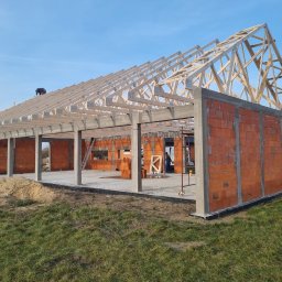 NOX INTERNATIONAL SPÓŁKA Z OGRANICZONĄ ODPOWIEDZIALNOŚCIĄ - Wznoszenie budynku z czerwonej cegły z drewnianą konstrukcją dachu w słoneczny dzień. Widoczny pracownik na dachu i stos cegieł wewnątrz budynku.