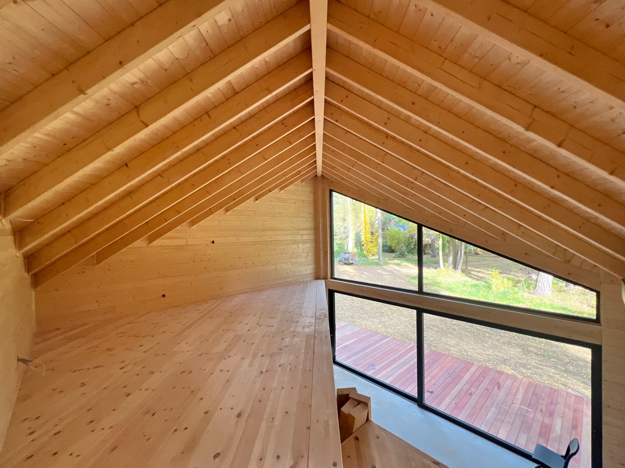 Wnętrze domu szkieletowego z drewnianą podłogą i ścianami, widok na zewnątrz przez duże okno. Charakterystyczna konstrukcja dachu z belek. Jasne, naturalne światło.