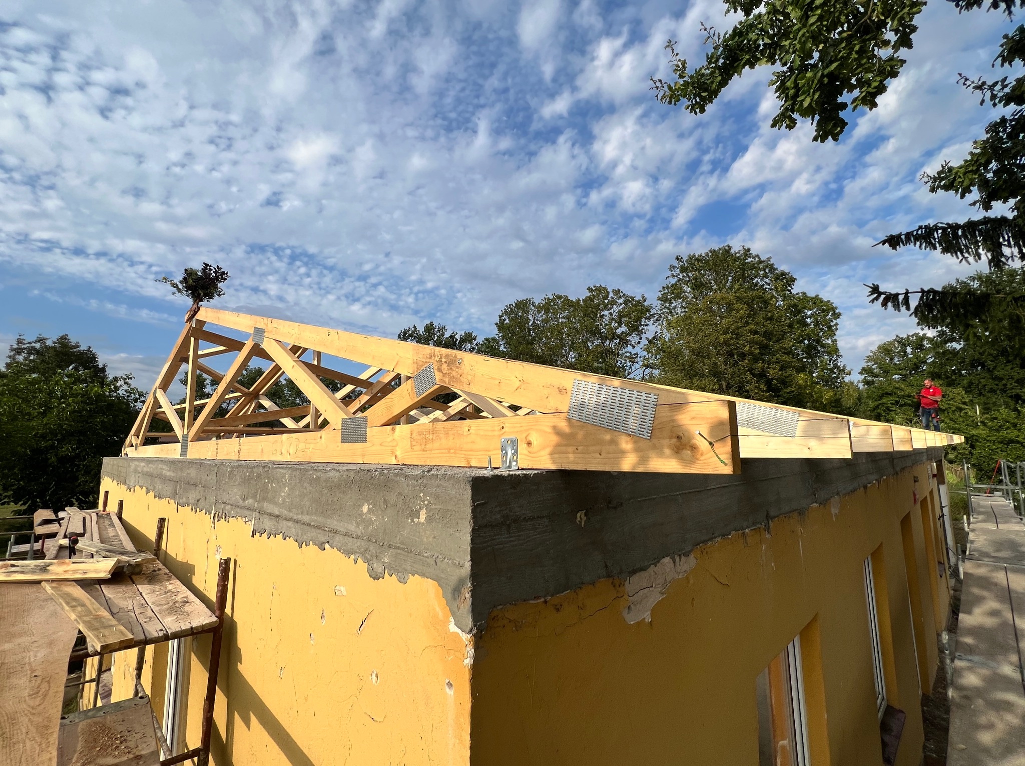 Wznoszenie drewnianej więźby dachowej na budynku z żółtą elewacją, widoczne elementy konstrukcyjne i metalowe łączniki, błękitne niebo w tle.