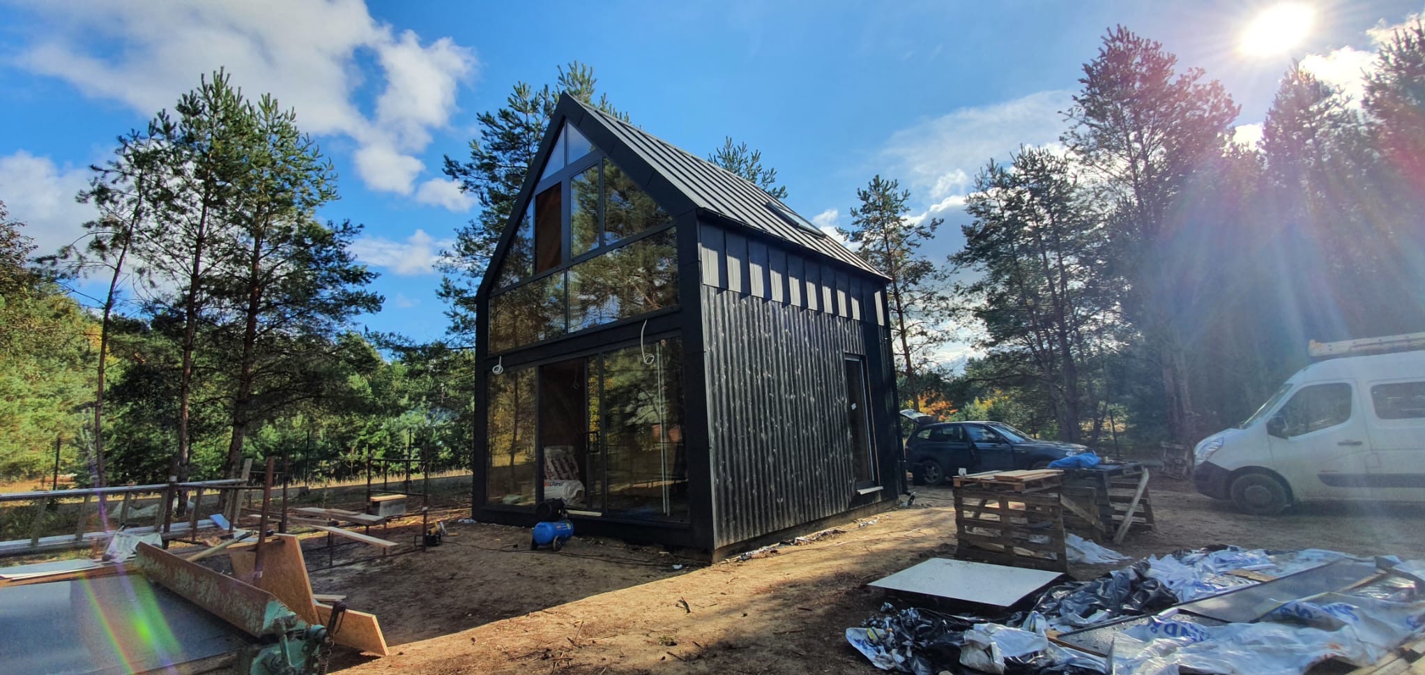 Nowoczesny, czarny dom modułowy z dużymi przeszkleniami, otoczony lasem. Widoczne elementy budowy i samochody w tle. Ujęcie w słoneczny dzień.