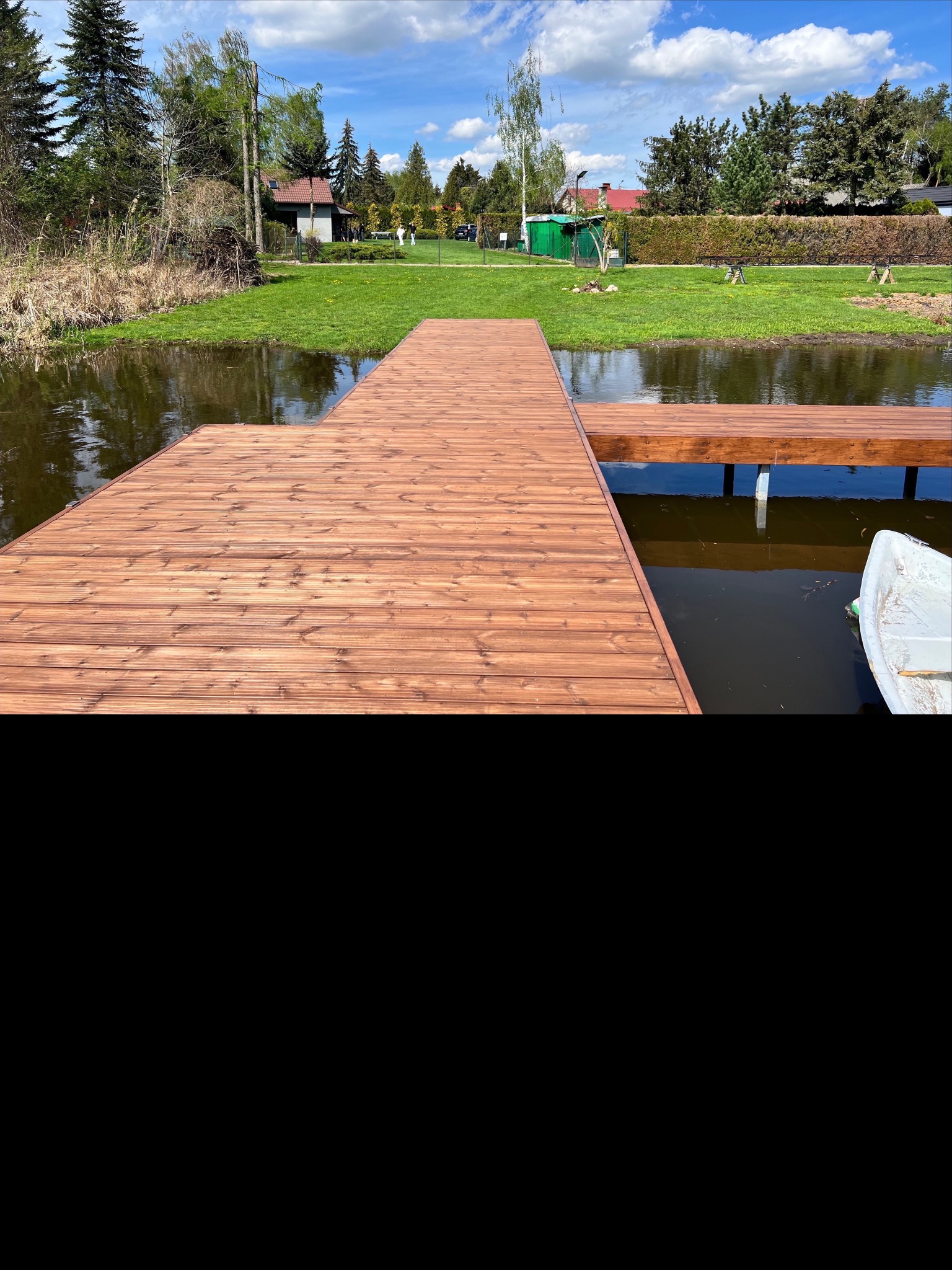 Drewniany pomost nad wodą w słoneczny dzień. Widoczne deski pomostu, woda, trawa, drzewa i budynki w tle. Część łódki przy pomoście. Jasne, naturalne światło.