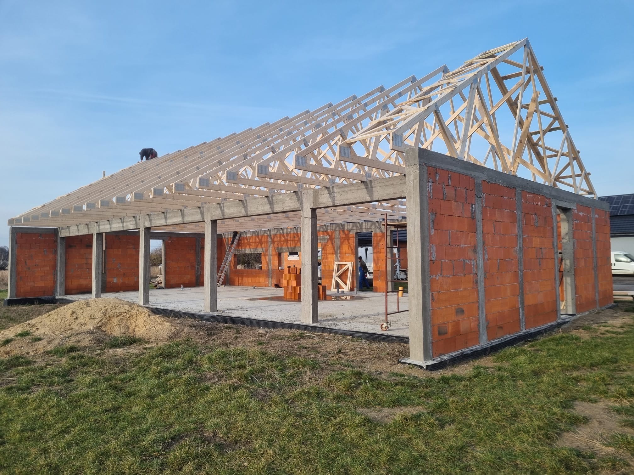 Wznoszenie budynku z czerwonej cegły z drewnianą konstrukcją dachu w słoneczny dzień. Widoczny pracownik na dachu i stos cegieł wewnątrz budynku.