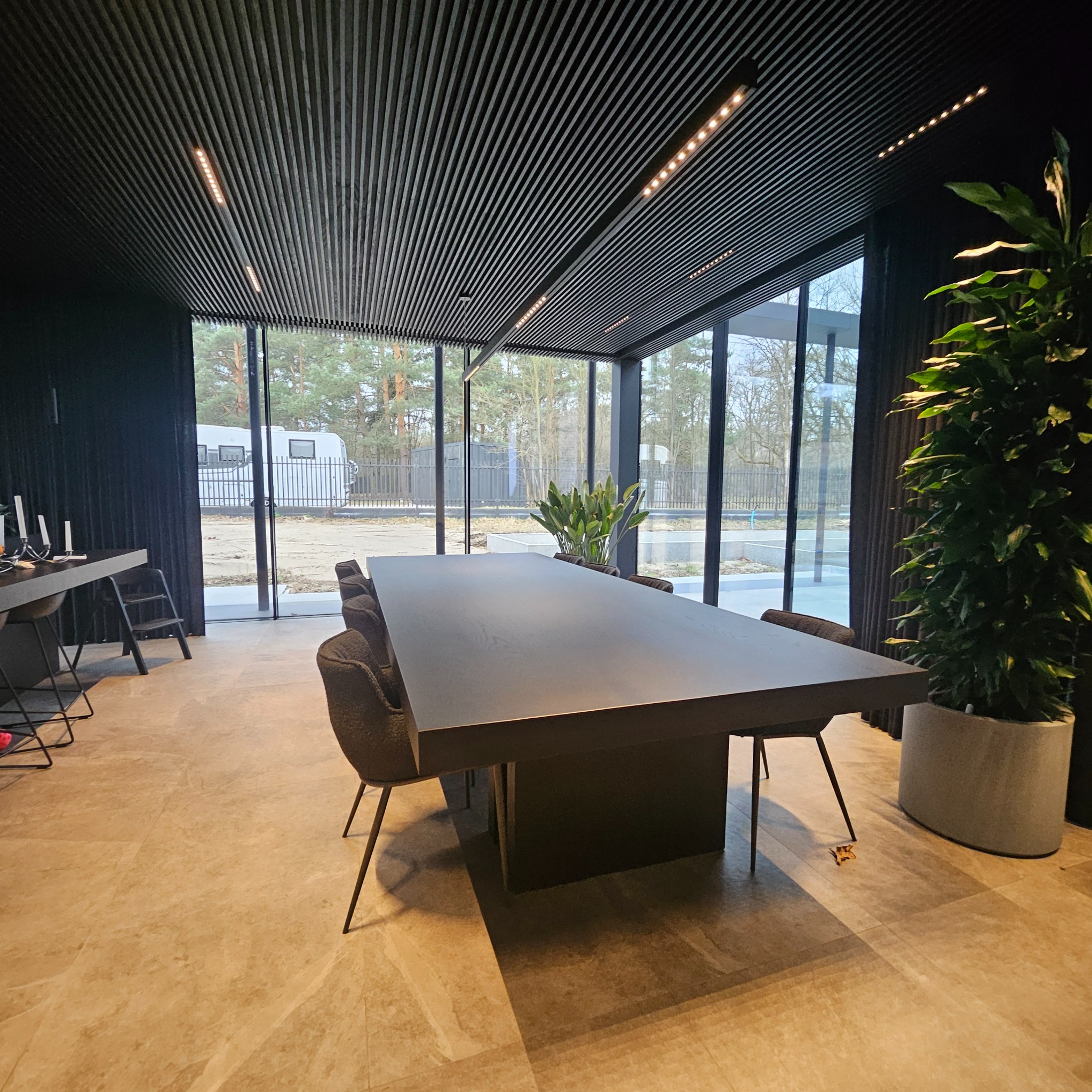 Nowoczesna jadalnia z dużym, ciemnym stołem i krzesłami na tle okien z widokiem na zewnątrz. Górę zdobi lamelowy sufit z oświetleniem punktowym.