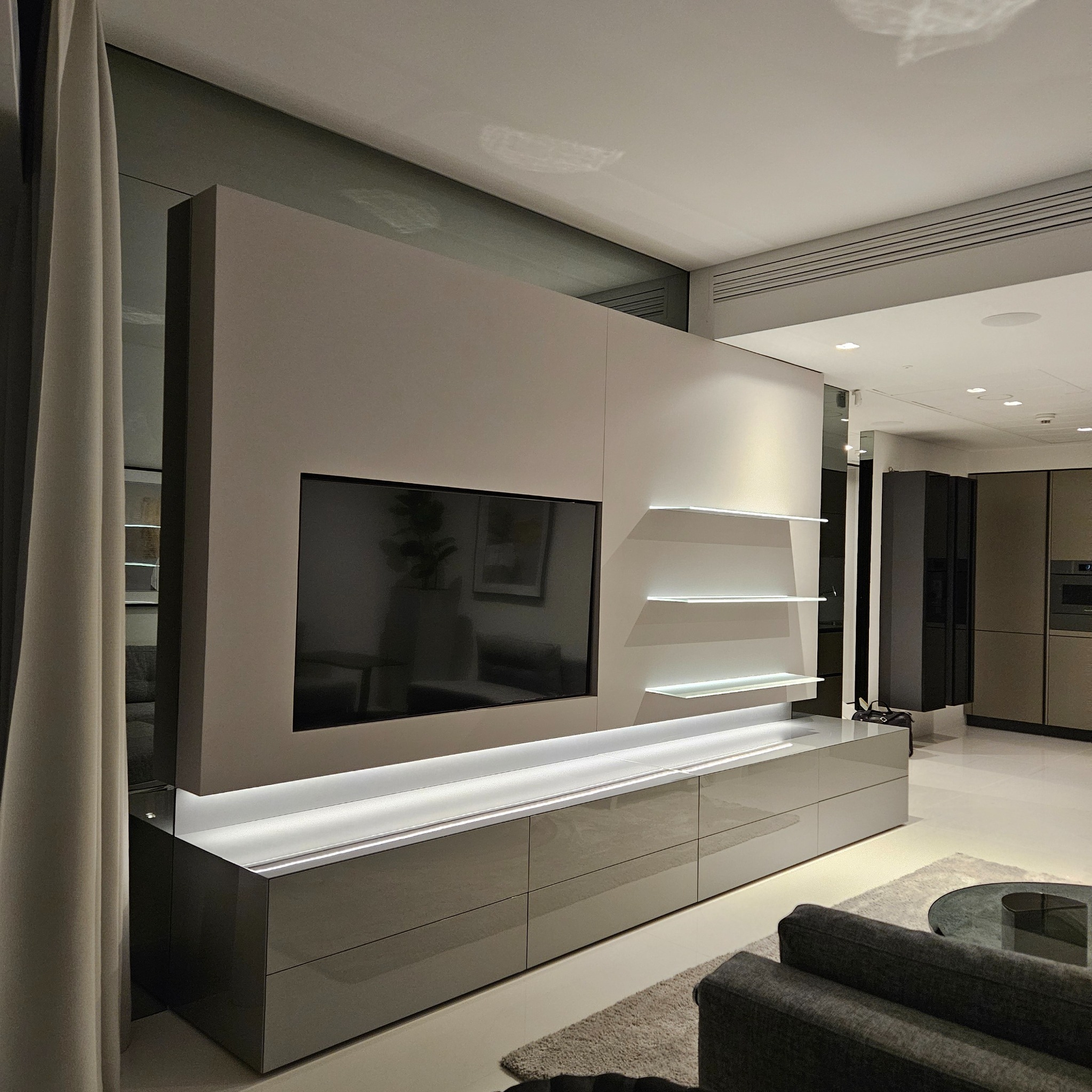 Nowoczesna zabudowa ściany z telewizorem i podświetlanymi półkami w salonie. Minimalistyczny design, gładkie fronty szafek, stonowana kolorystyka, widok na kuchnię.