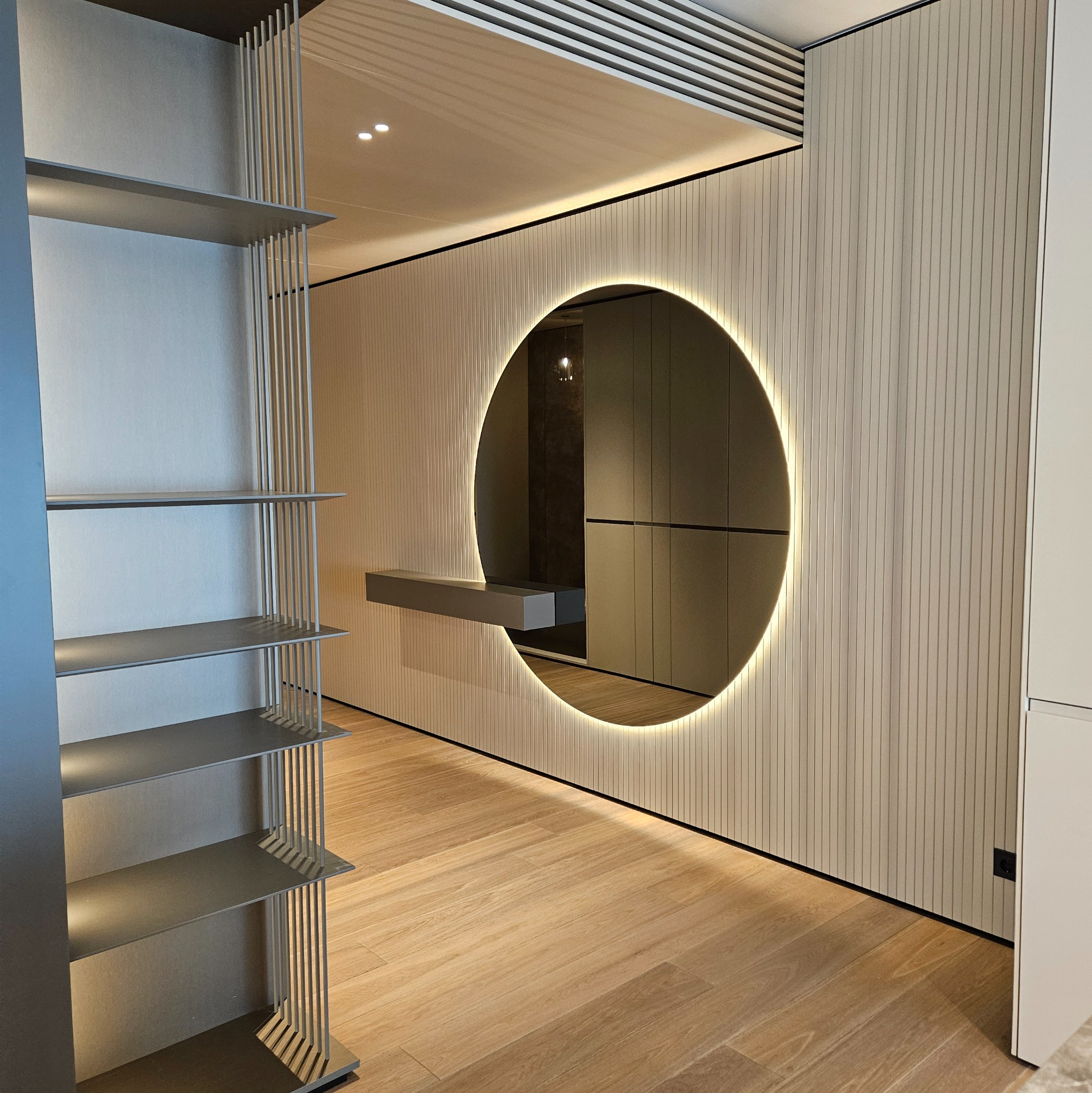 Nowoczesna garderoba z podświetlanym lustrem w okrągłej ramie i półkami z metalowymi prętami, drewniana podłoga, minimalistyczny design.