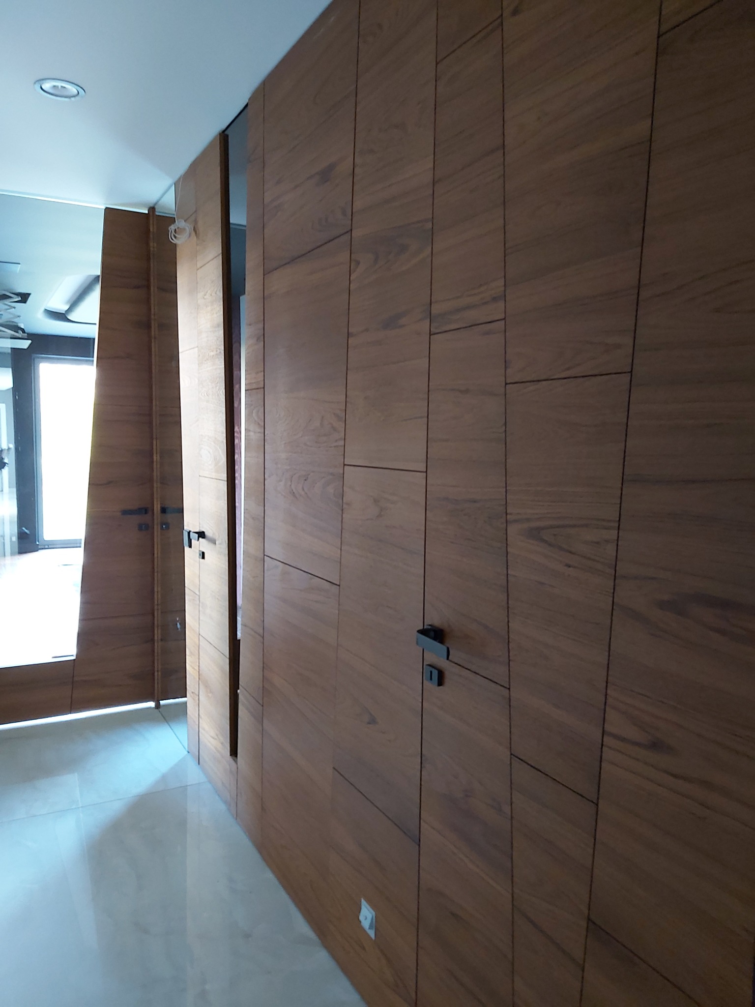 Drewniana zabudowa wnęki z ukrytymi drzwiami i szafkami, minimalistyczne czarne uchwyty, oświetlone wnętrze, jasna podłoga, nowoczesny design.