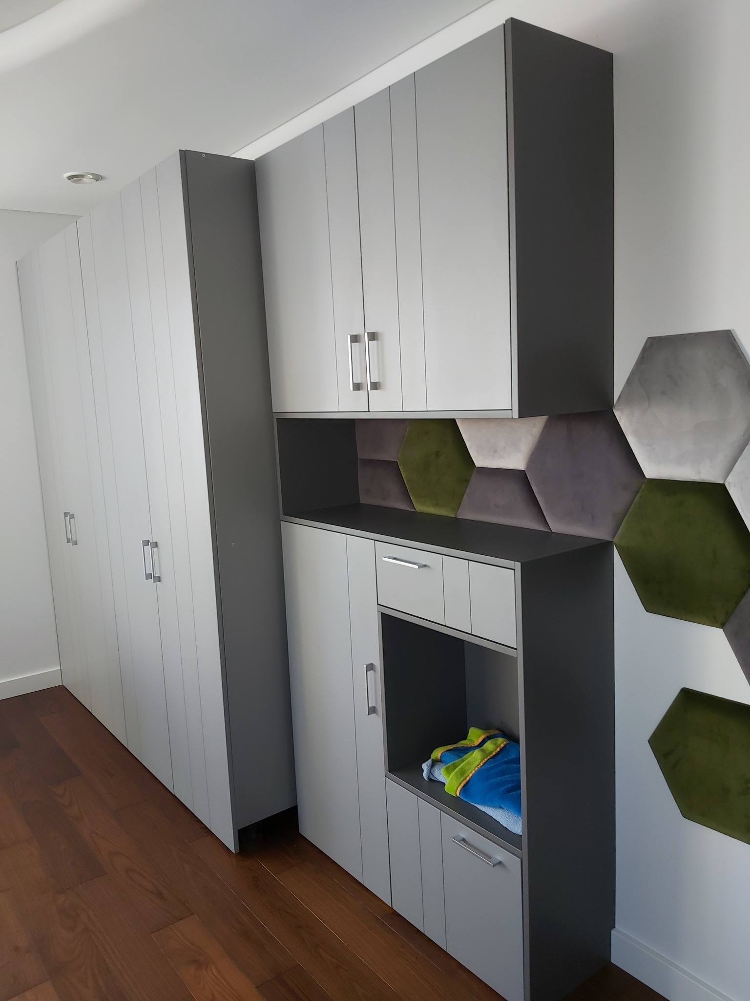 Szara zabudowa meblowa z szafkami i półkami, uzupełniona o heksagonalne panele dekoracyjne w odcieniach szarości i zieleni. Uwagę zwraca nowoczesny design i funkcjonalność.