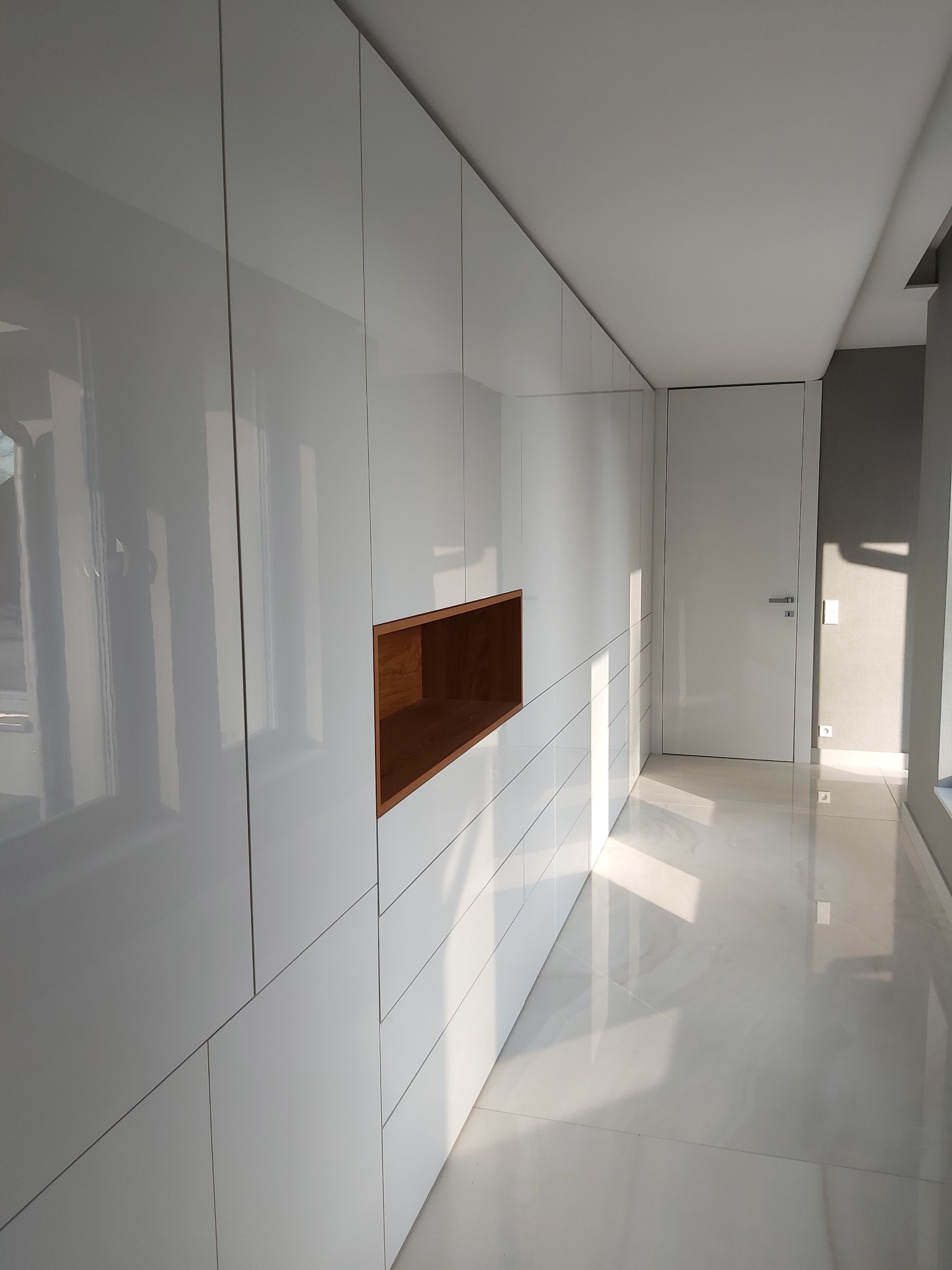 Biała, lakierowana szafa wnękowa z drewnianą wnęką, w nowoczesnym korytarzu z połyskującą podłogą. Minimalistyczny design i jasne, naturalne światło.