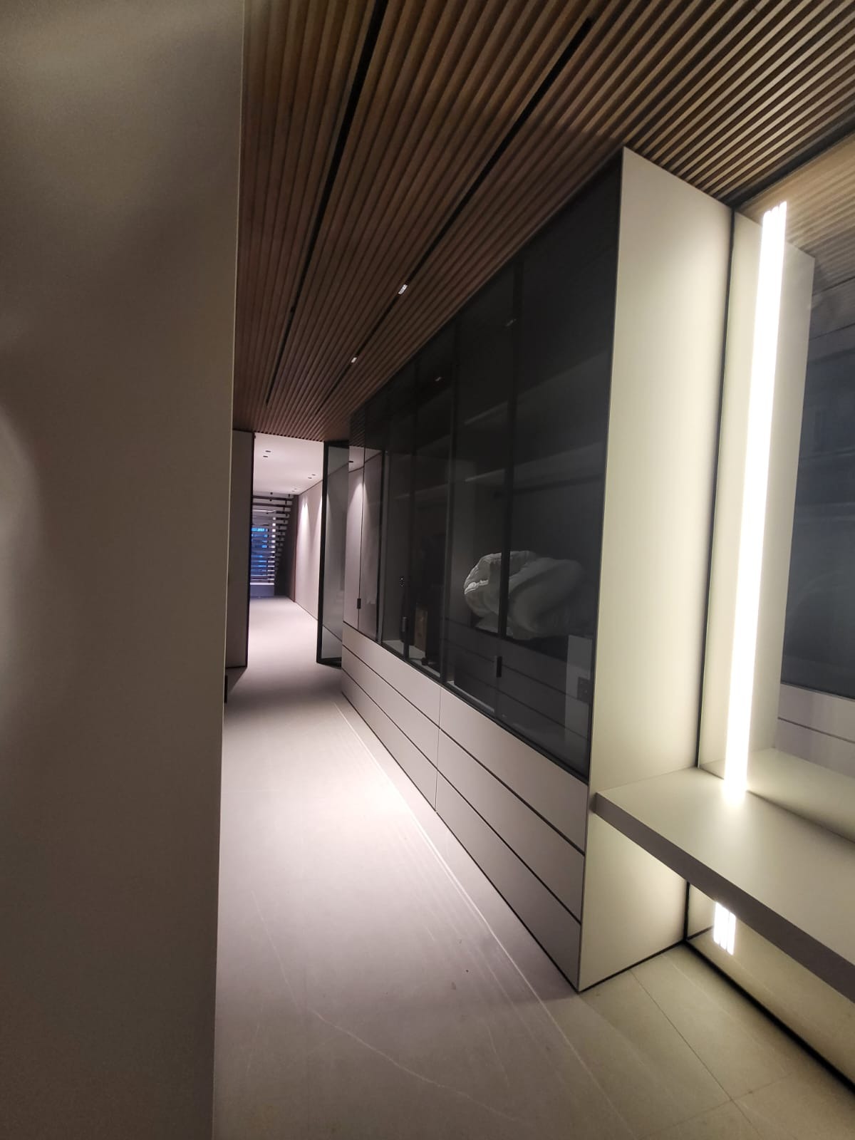 Długa, nowoczesna garderoba z podświetlanymi szafami z ciemnymi szybami i drewnianym sufitem lamelowym, widziana z perspektywy korytarza.