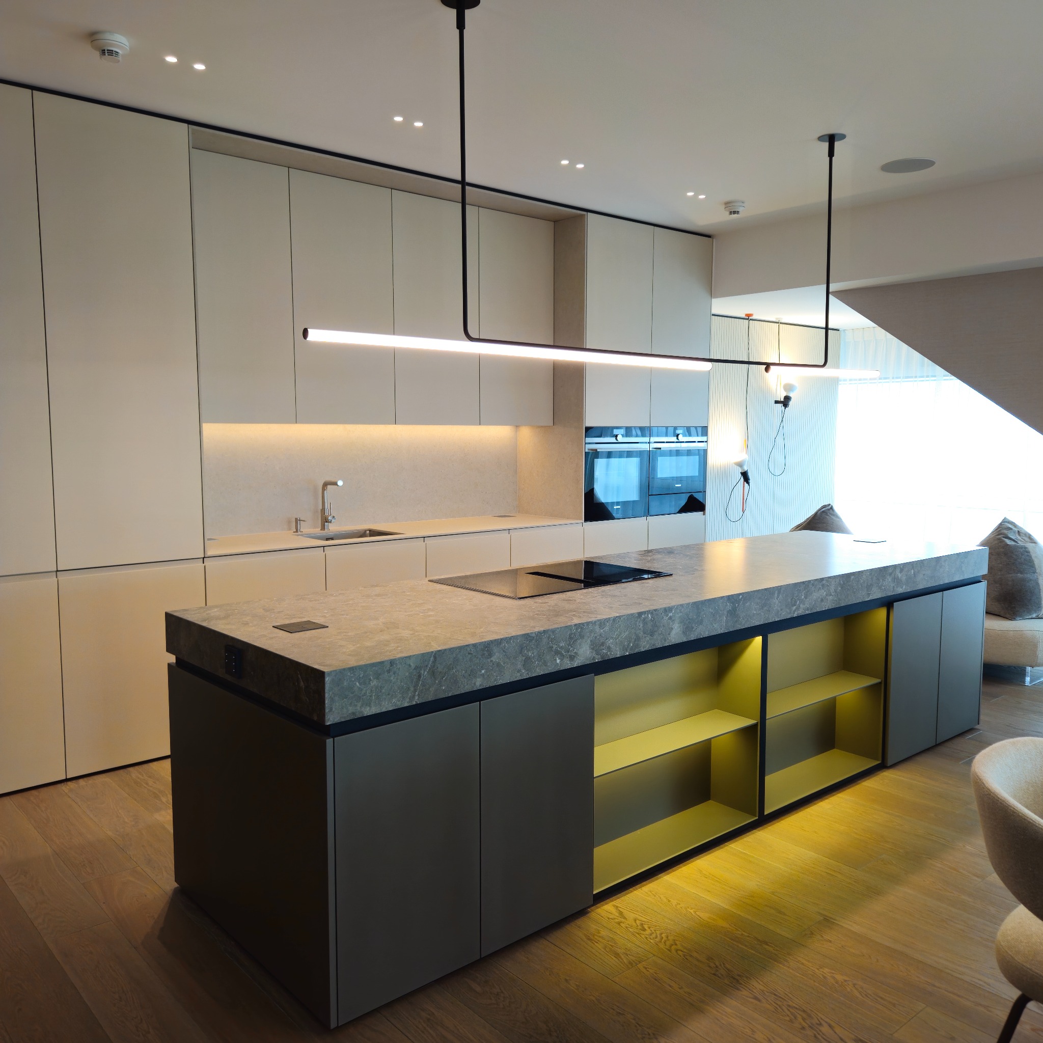 Nowoczesna kuchnia z wyspą i oświetleniem LED. Blat z szarego kamienia, fronty szafek w odcieniach beżu i szarości. Żółte półki w wyspie dodają akcentu kolorystycznego.