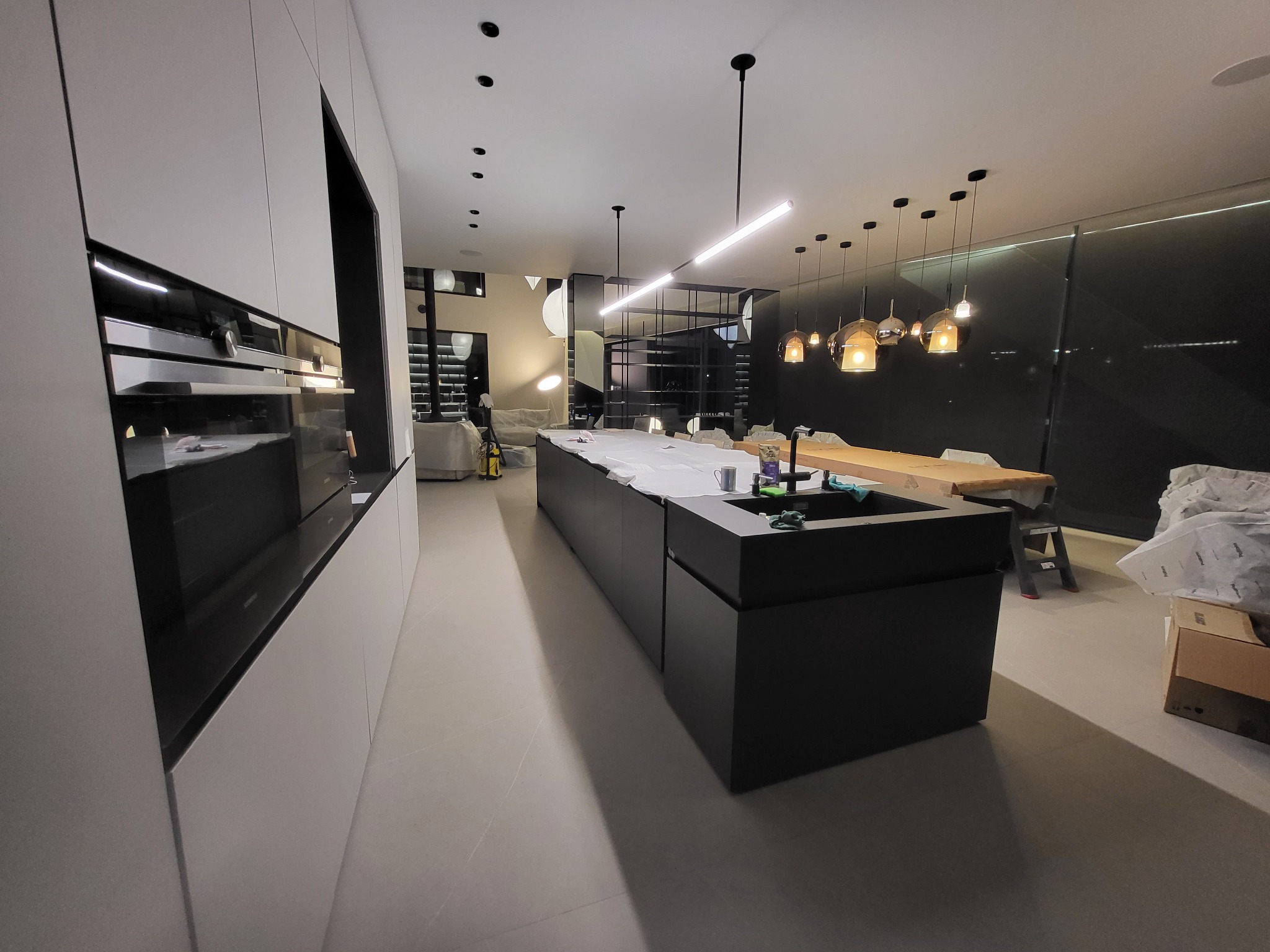 Nowoczesna kuchnia z wyspą w trakcie montażu. Widoczne minimalistyczne szafki w kolorze białym i czarnym, designerskie oświetlenie i sprzęt AGD.
