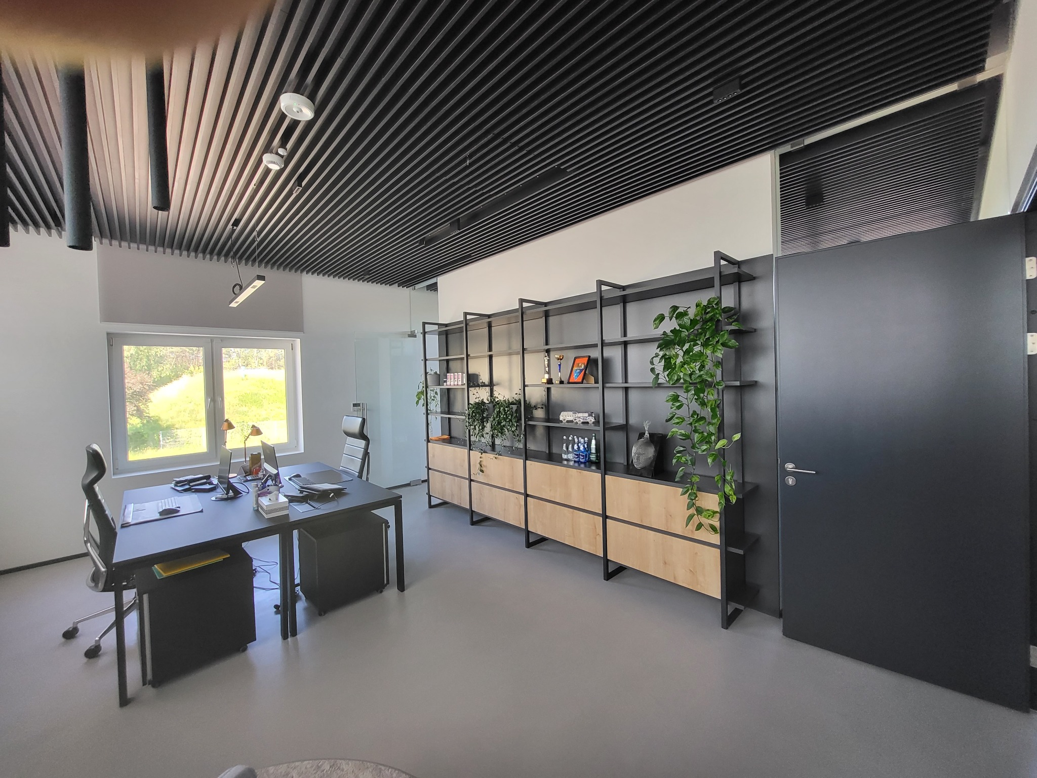 Nowoczesne biuro z czarnymi meblami: biurko, regał z drewnianymi akcentami i roślinami. Sufit z lamelami, widok na zewnątrz przez okno. Minimalistyczny design.