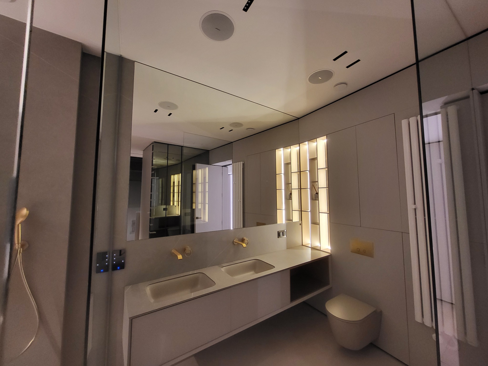 Nowoczesna łazienka z podwójną umywalką, złotą armaturą i oświetleniem LED. W lustrze odbija się kabina prysznicowa i fragment garderoby.