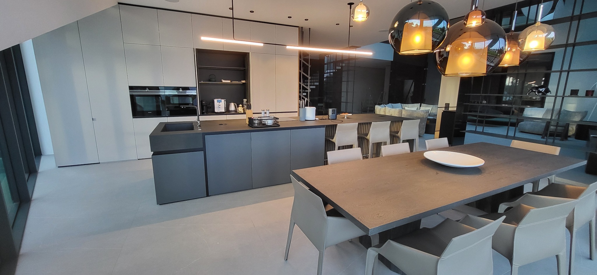 Nowoczesna kuchnia z wyspą i stołem jadalnym, minimalistyczny design, szaro-biała kolorystyka, oświetlenie LED, widok na salon z przeszkloną ścianą.