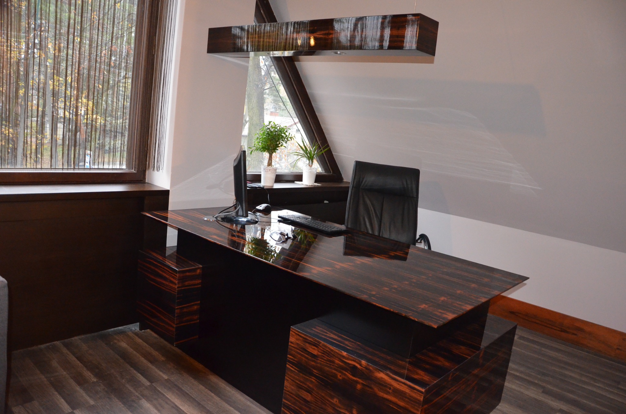 Eleganckie biuro z ciemnego drewna z widokiem na okno. Na biurku komputer, klawiatura, okulary i doniczki z roślinami. Nad biurkiem designerska lampa.