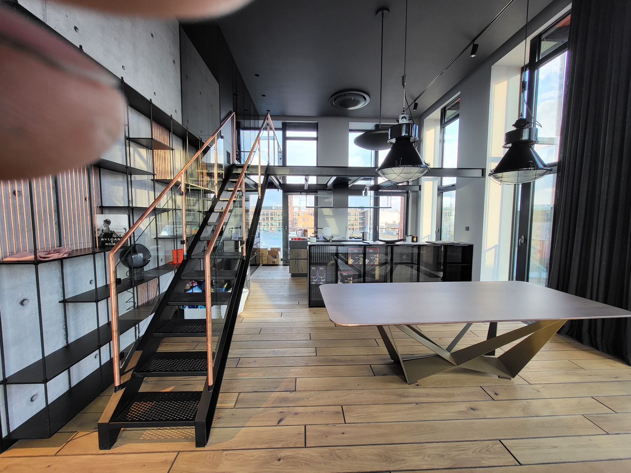 Nowoczesne wnętrze loftu z otwartą przestrzenią: schody z metalu i szkła, stół z oryginalną podstawą i widok na miasto przez duże okna. Industrialny design.